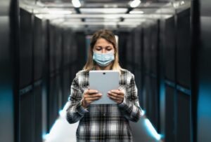 Female informatic engineer working inside server room database while wearing face mask